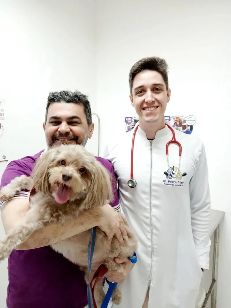 Tutor sorridente com seu cão no colo ao lado do Dr. Pedro Stipp, de estetoscópio, depois da consulta